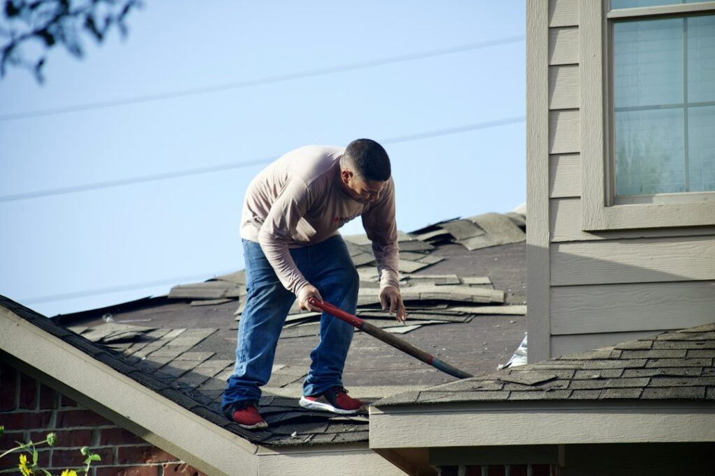 A homeowner standing on a ladder clearing leaves from rain gutters on a two-story house.
