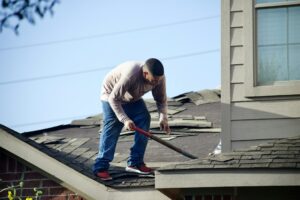 A homeowner standing on a ladder clearing leaves from rain gutters on a two-story house.